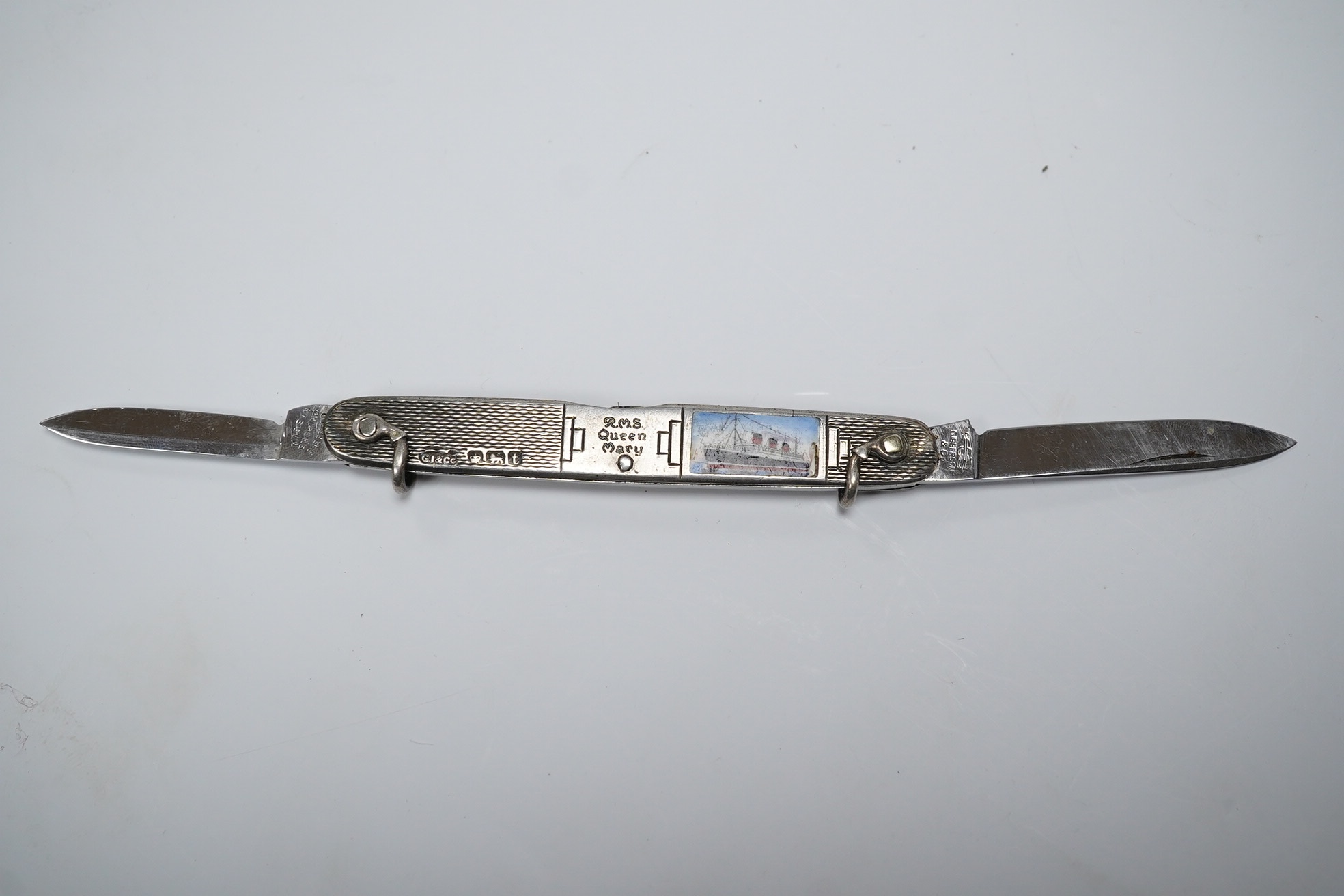 A George V engine turned silver and enamel pocket knife, decorated with R.M.S Queen Mary, the blades lifted by the handles, George Ibberson & Co, Sheffield, 1936, 76mm. Condition - poor to fair